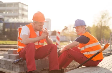 two men in hardhats and safety vests sit down for a cigarette