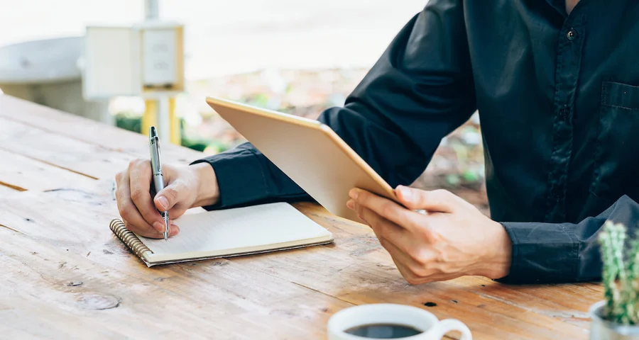 person taking notes on paper while holding tablet