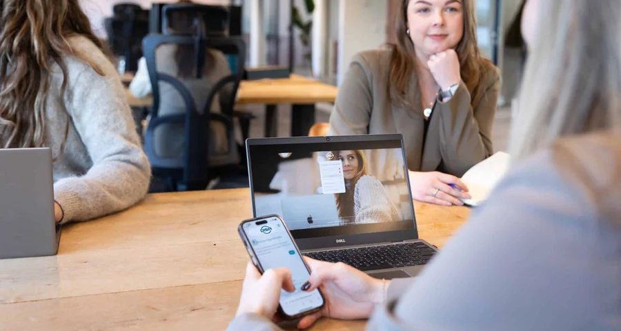 Teamet vid bordet samarbetar med hjälp av en compliance-app för smartphones och ett videosamtal på en bärbar dator, vilket visar på enhetlig integritet.