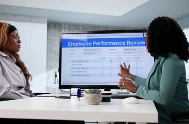 Employee Performance Review, an employee sits at a desk with their manager reviewing their performance on a computer screen.