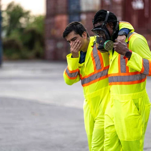 industrial workers in high-visibility suits assist each other after a hazardous incident near a shipping container. Emphasizing occupational safety, emergency response, and risk management