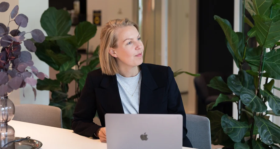 Kvinna i kavaj som sitter framför dator i kontorsmiljö och tittar upp till höger.