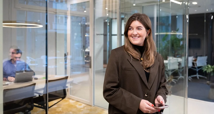 vrouw in een bruin jasje staat op kantoor met haar telefoon in de hand en lacht